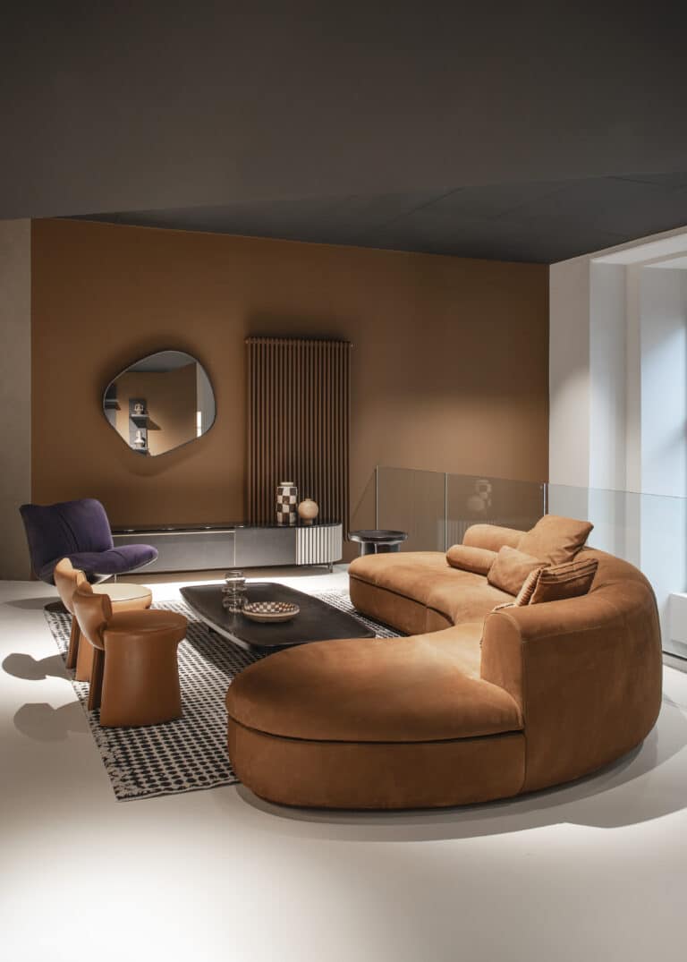 Showroom zeigt einen Wohnzimmertypischen Aufbau mit Sofa in einemdunkel orangenen Ton, dazu ein tiefer Couchtisch und weitere Sitzmöglichkeiten in form von Sesseln. Im Hintergrund sieht man einen runden Spiegel an der Wand hängen.